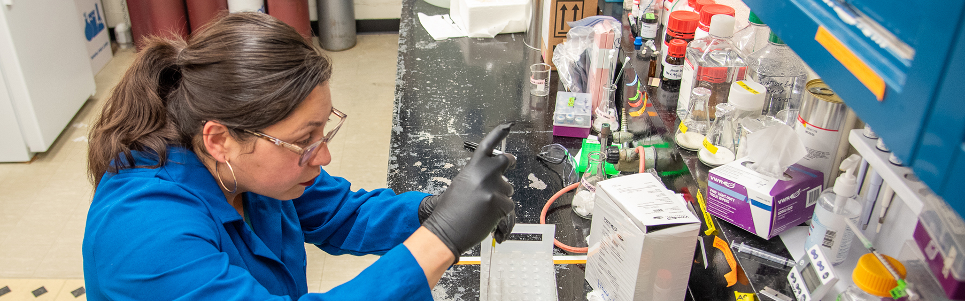 a woman in a lab with lab equipment on the table.  She is putting material into a vial.